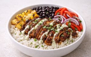 Caribbean jerk chicken rice bowl with coconut rice, charred pineapple, black beans, red peppers, and lime-cilantro crema garnished with fresh cilantro