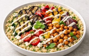 Colorful Mediterranean veggie bowl with grilled zucchini, eggplant, peppers, lemon-herb quinoa, crispy chickpeas, feta cheese, and drizzled tahini-yogurt sauce in a white bowl
