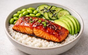 Miso glazed salmon rice bowl with sushi rice, edamame, pickled cucumbers, avocado, and nori garnished with sesame seeds and scallions