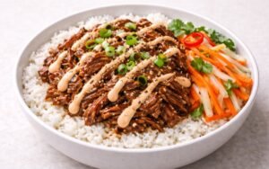 Pulled pork rice bowl with glazed shredded pork, pickled vegetables, sriracha mayo, and sesame seeds over jasmine rice