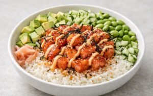 Colorful spicy salmon sushi bowl with seared salmon cubes, sliced avocado, cucumber, edamame, and drizzled spicy mayo over white sushi rice in a ceramic bowl