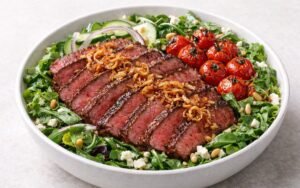 Steak salad bowl with sliced sirloin, mixed greens, roasted cherry tomatoes, feta cheese, and garlic vinaigrette in a white bowl