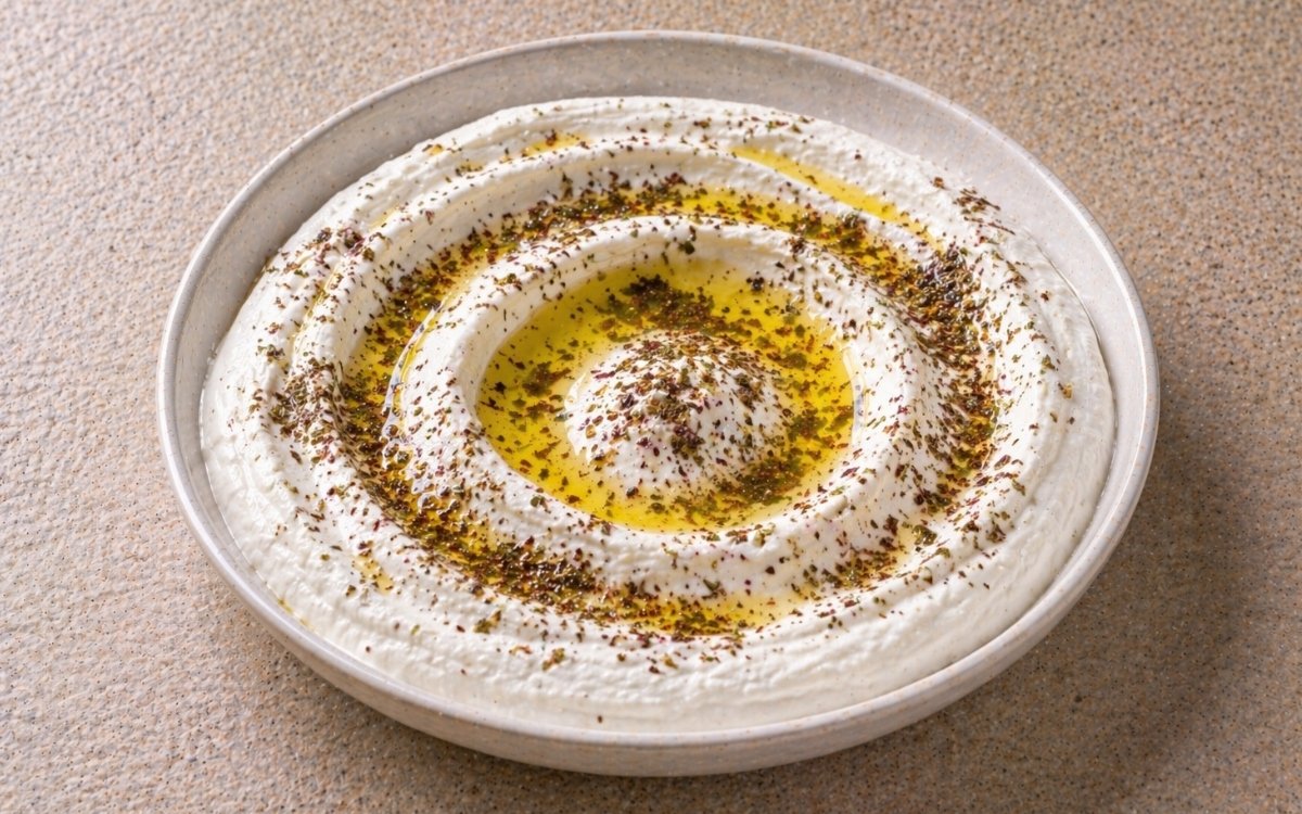 Authentic labneh in a wide white bowl showing creamy white surface with olive oil well, generous za'atar garnish, and warm pita bread beside it on marble surface