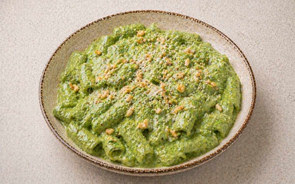 Creamy spinach herb green sauce pasta in a wide white bowl showing vivid green sauce coating rigatoni with toasted pine nuts, extra Parmigiano, and fresh herbs on marble surface