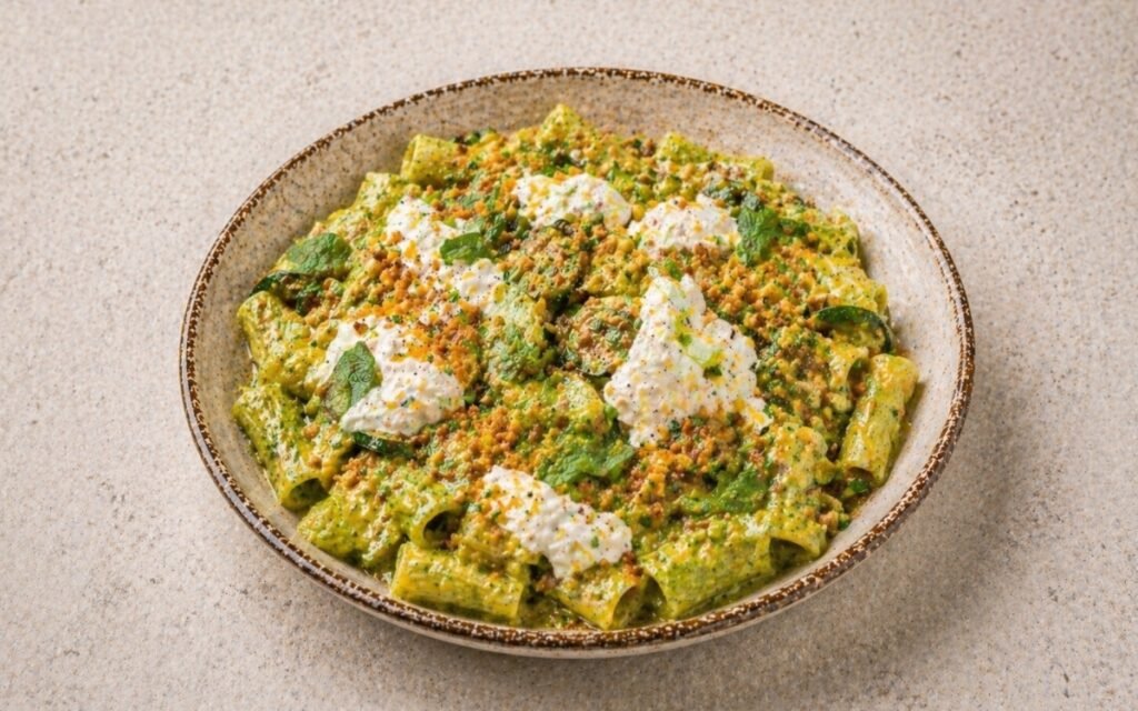 Creamy zucchini pasta in a wide shallow white bowl showing vivid green sauce coating rigatoni with intact caramelised zucchini pieces, torn burrata with cream pooling, lemon breadcrumbs, and fresh basil on marble surface