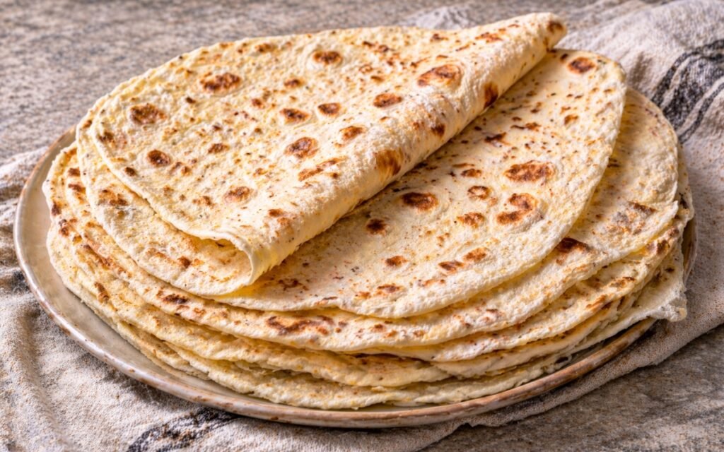 Stack of homemade lavash flatbread showing golden blister spots and soft pliable texture on a cloth-lined wooden board with fresh herbs and dipping sauce beside it
