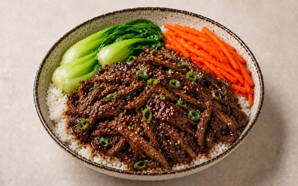 Korean-style braised beef short rib rice bowl in a wide shallow bowl showing glossy shredded short rib over jasmine rice with blanched bok choy, julienned carrot, scallions, sesame seeds, and sauce drizzled over on marble surface