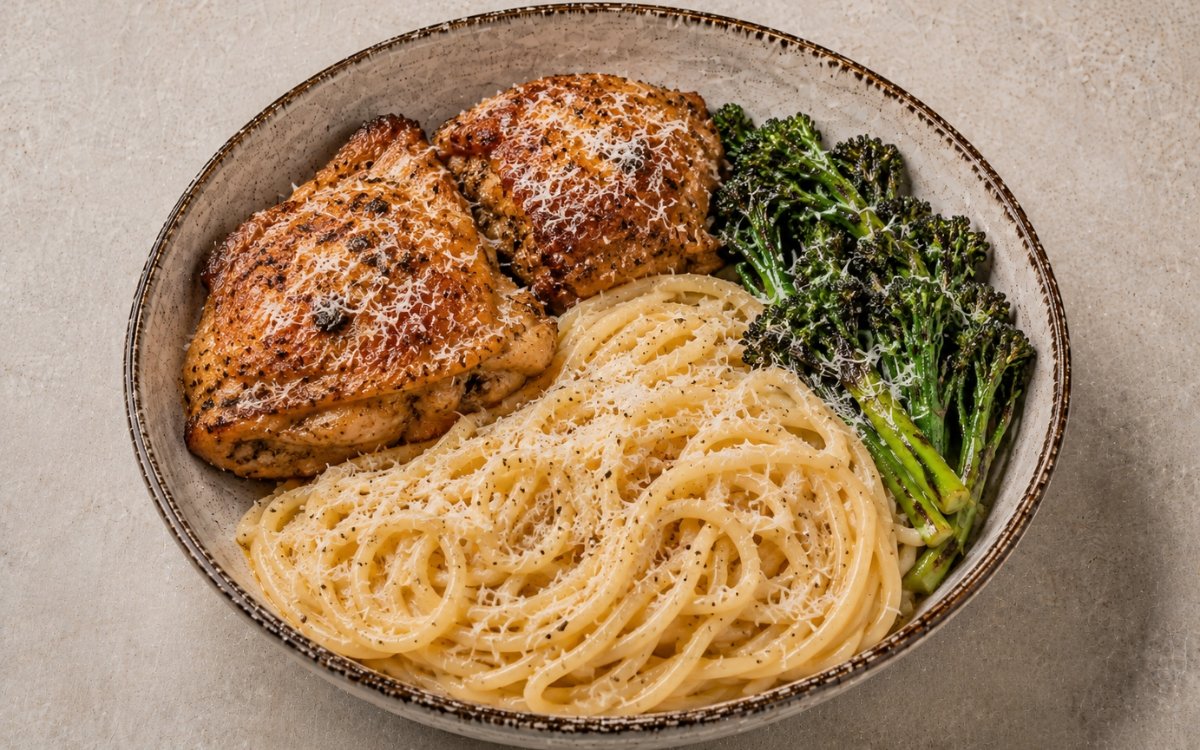 Oven baked chicken thighs with cheesy spaghetti in a wide shallow bowl showing two deeply golden crispy chicken thighs alongside cacio e pepe spaghetti, charred broccolini, and a heavy grating of Parmesan on marble surface