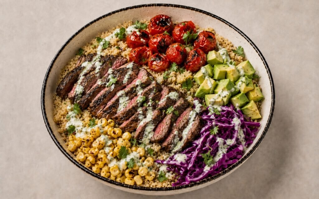 Light skirt steak grain bowl in a wide shallow bowl showing thinly sliced medium-rare spiced skirt steak over quinoa with charred cherry tomatoes, corn, diced avocado, shredded red cabbage, and cilantro-lime dressing