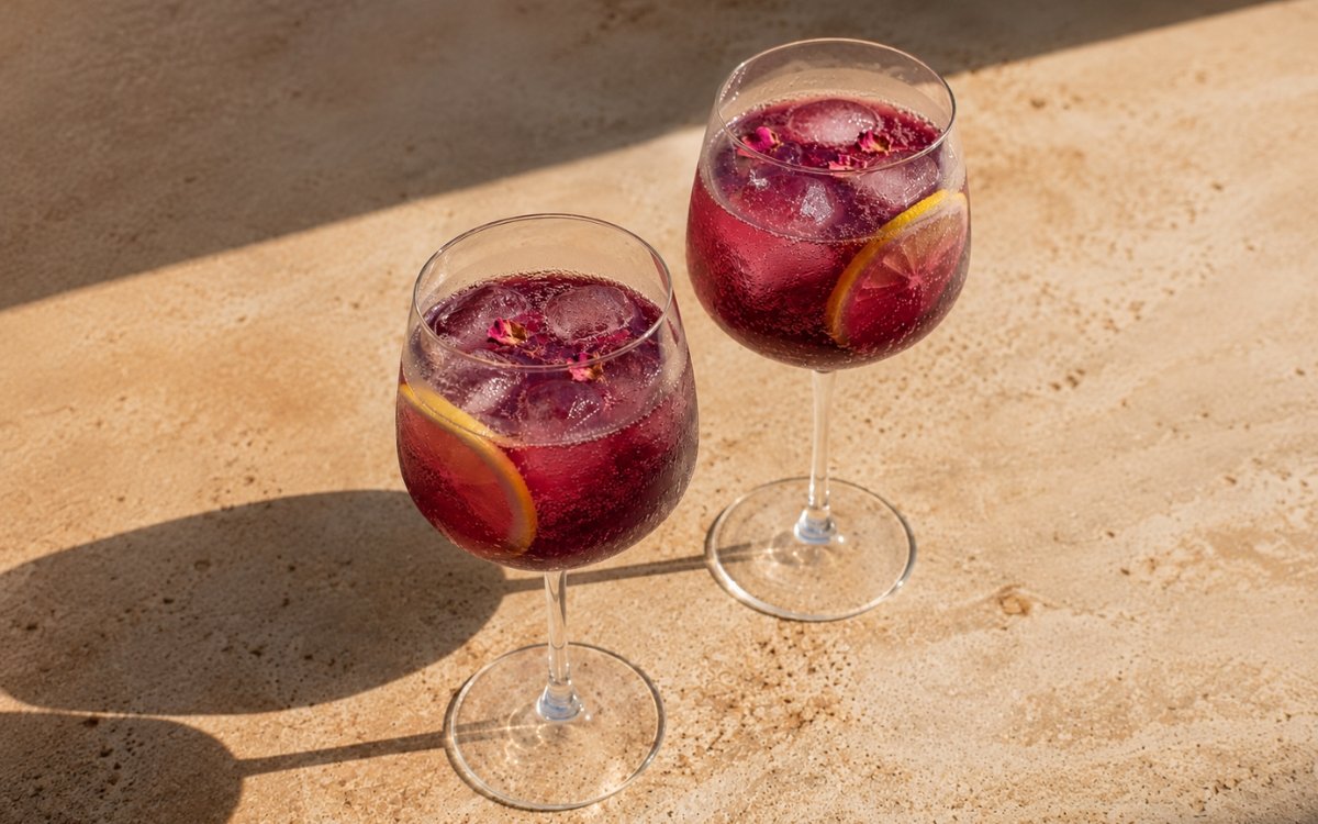 Blackcurrant rose fizz mocktail in a large wine glass showing vivid deep purple sparkling drink over ice with dried rose petals scattered on the surface and a thin lemon slice against the glass on marble surface
