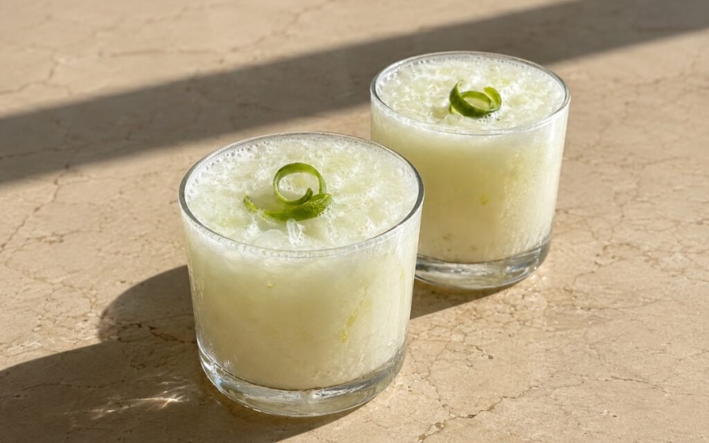 Brazilian lemonade in short rocks glasses with creamy opaque white-green liquid, crushed ice and lime peel curl on a warm beige marble surface