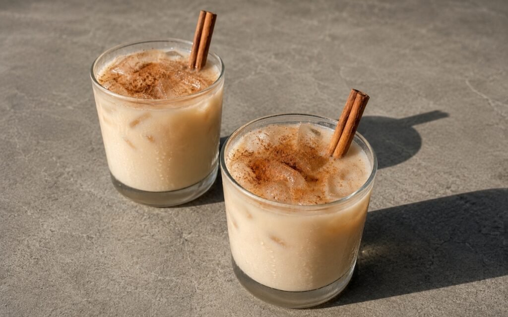 Chocolate cinnamon horchata in short rocks glasses with creamy beige liquid, ice cubes, cinnamon powder dusting and cinnamon stick on a warm grey stone surface
