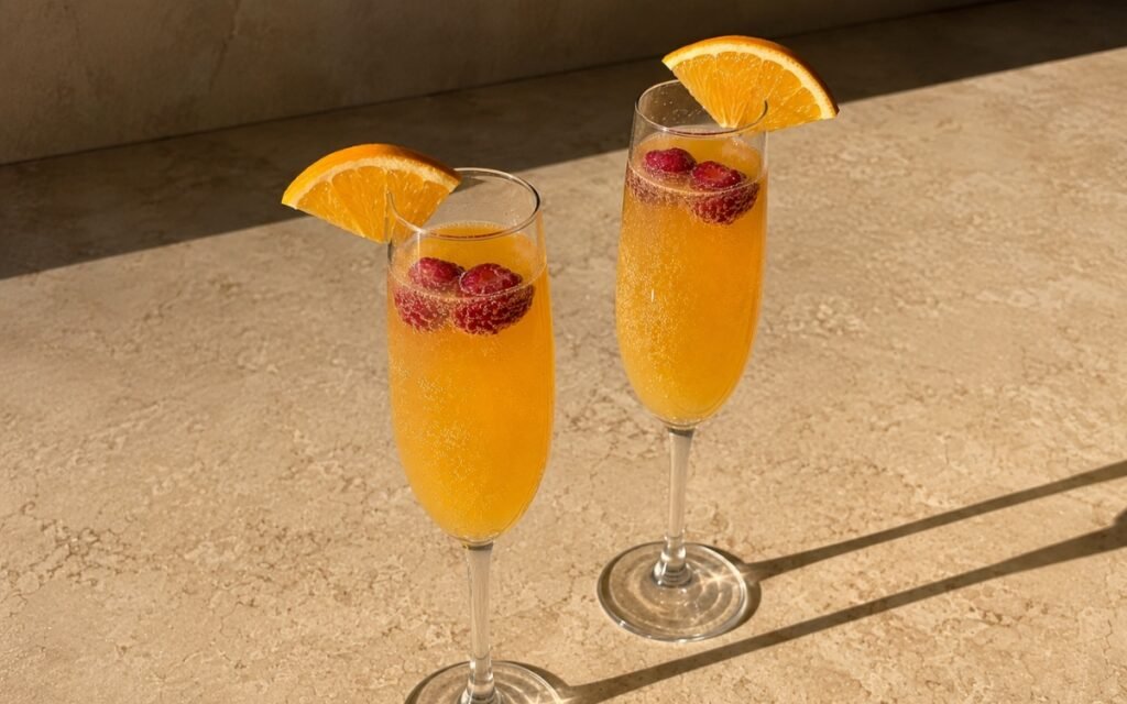 Classic mimosa mocktail in champagne flute glasses with golden-orange sparkling liquid, orange wedge on the rim and fresh raspberries floating on a warm marble surface