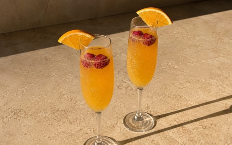 Classic mimosa mocktail in champagne flute glasses with golden-orange sparkling liquid, orange wedge on the rim and fresh raspberries floating on a warm marble surface