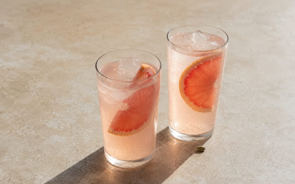 Grapefruit cardamom tonic mocktail in a tall glass showing pale blush sparkling drink over ice with a thick grapefruit slice tucked into the drink on marble surface