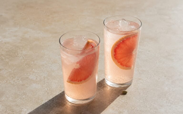 Grapefruit cardamom tonic mocktail in a tall glass showing pale blush sparkling drink over ice with a thick grapefruit slice tucked into the drink on marble surface
