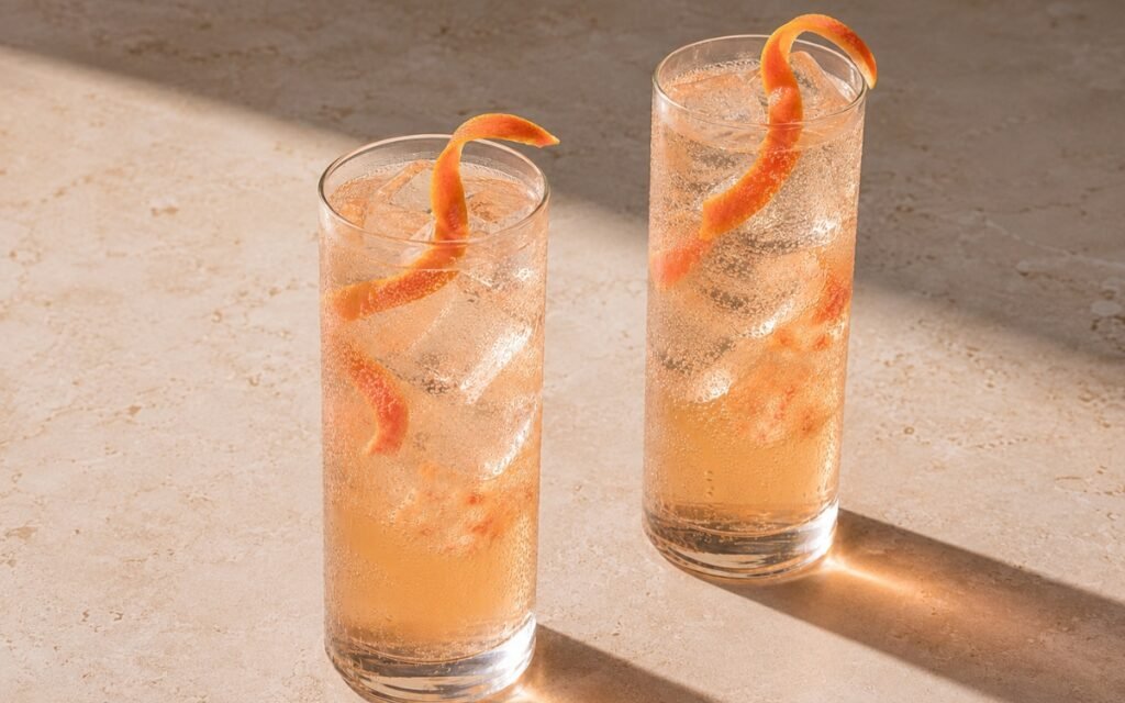 Grapefruit tonic mocktail in tall highball glasses with pale blush-pink sparkling liquid, ice cubes and grapefruit peel curl draped from the rim on a light warm stone surface