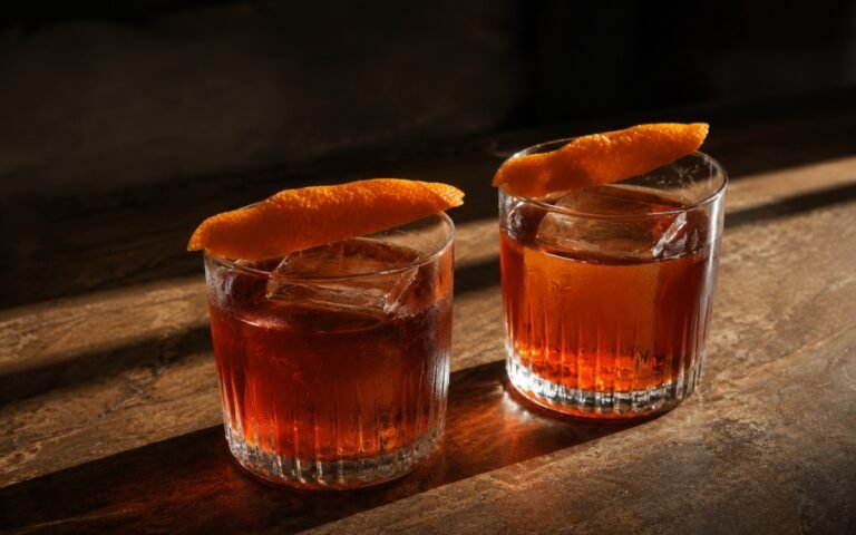 Negroni sbagliato style bitter orange mocktail in an old fashioned glass showing deep amber drink over a single large clear ice cube with an expressed orange peel draped across the rim on marble surface