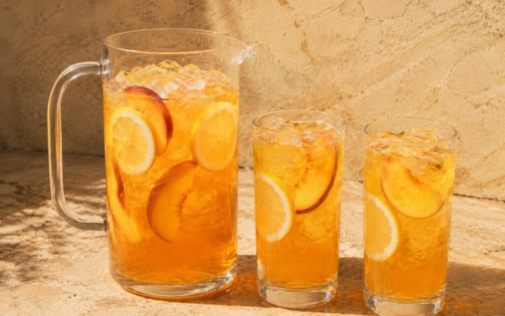 Large peach lemonade pitcher showing golden-amber still drink with lemon slices and fresh peach slices visible throughout on marble surface
