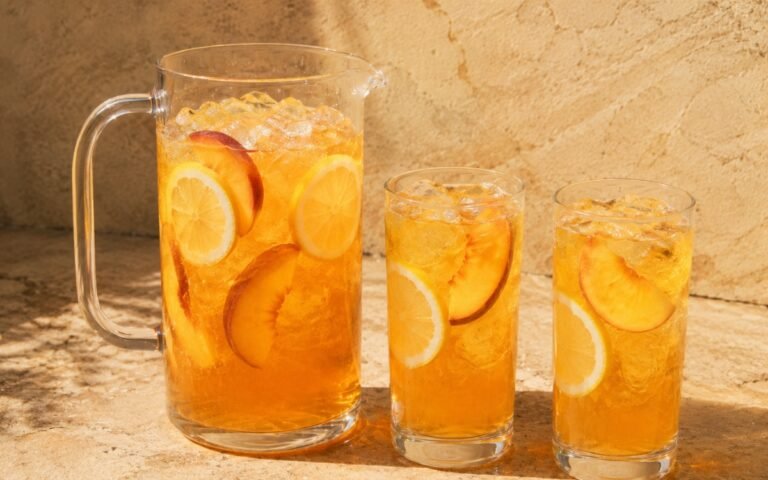Large peach lemonade pitcher showing golden-amber still drink with lemon slices and fresh peach slices visible throughout on marble surface
