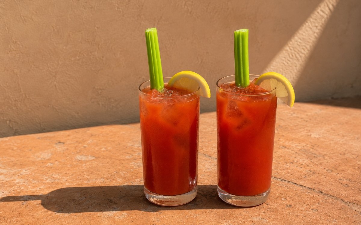 Tomato basil virgin mary mocktail in a tall glass showing vivid deep red still drink over ice with a celery stalk, lemon wheel, and generous cracked black pepper on marble surface