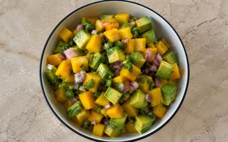 Avocado and mango salsa in a white bowl showing orange mango cubes, green avocado, red onion, fresh cilantro, and lime wedges on marble surface