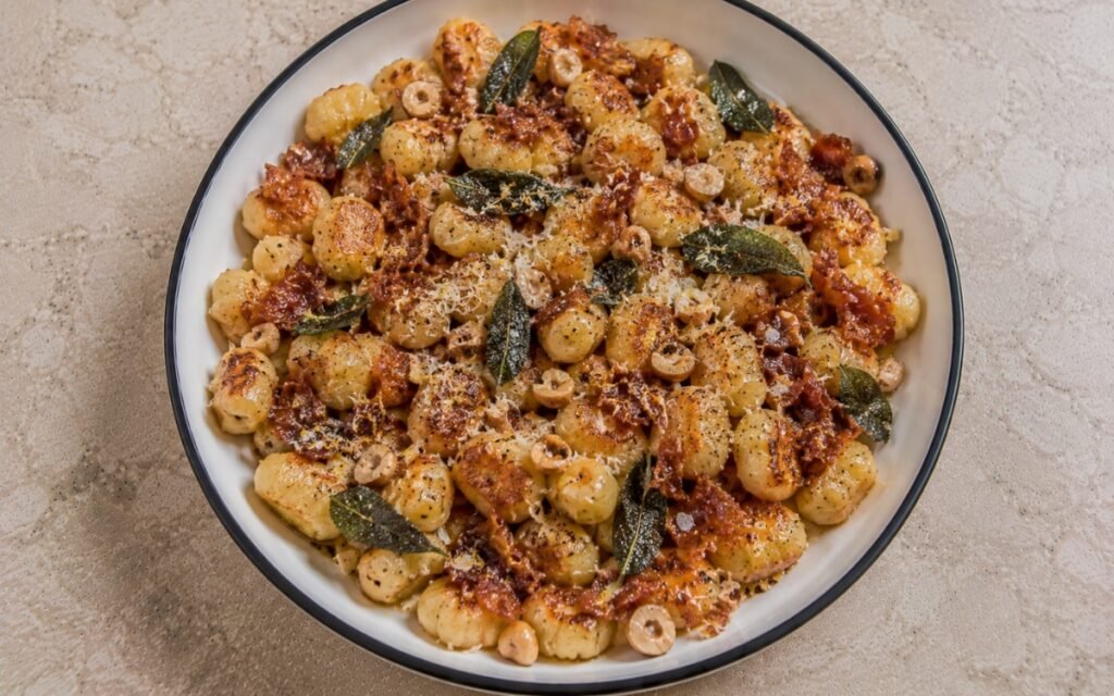Brown butter sage gnocchi with golden pillowy gnocchi in amber brown butter sauce, crispy fried sage leaves, crispy prosciutto, toasted hazelnuts, lemon zest and Parmigiano-Reggiano in a white ceramic bowl with black rim