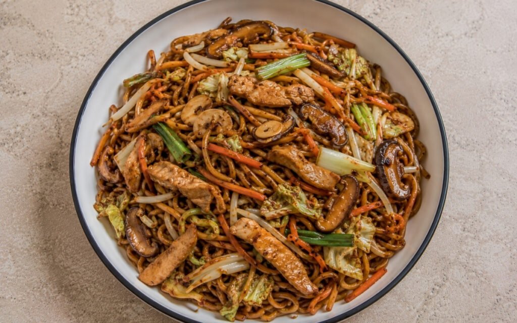 Chicken chow mein with fresh Hong Kong egg noodles in glossy dark soy-oyster sauce, sliced chicken thighs, napa cabbage, bean sprouts, shiitake, julienned carrots, scallions and julienned ginger in a white ceramic bowl with black rim