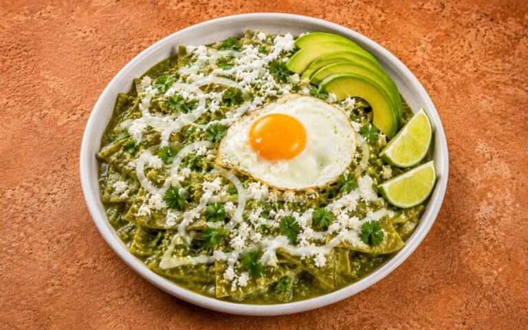 Bowl of chilaquiles verdes showing tortilla chips coated in vivid green salsa verde, topped with a fried egg with bright orange yolk, fanned avocado, cotija, onion rings, and crema drizzle on marble surface