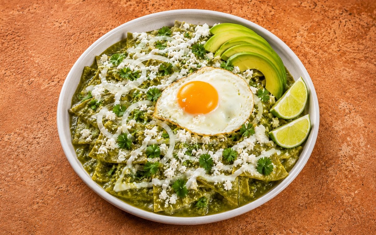 Bowl of chilaquiles verdes showing tortilla chips coated in vivid green salsa verde, topped with a fried egg with bright orange yolk, fanned avocado, cotija, onion rings, and crema drizzle on marble surface