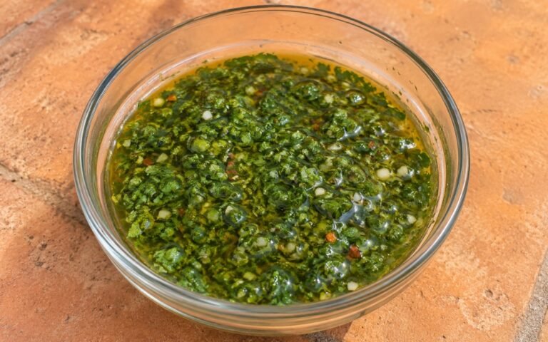 Bright green chimichurri sauce in a white bowl with fresh parsley and garlic cloves beside it