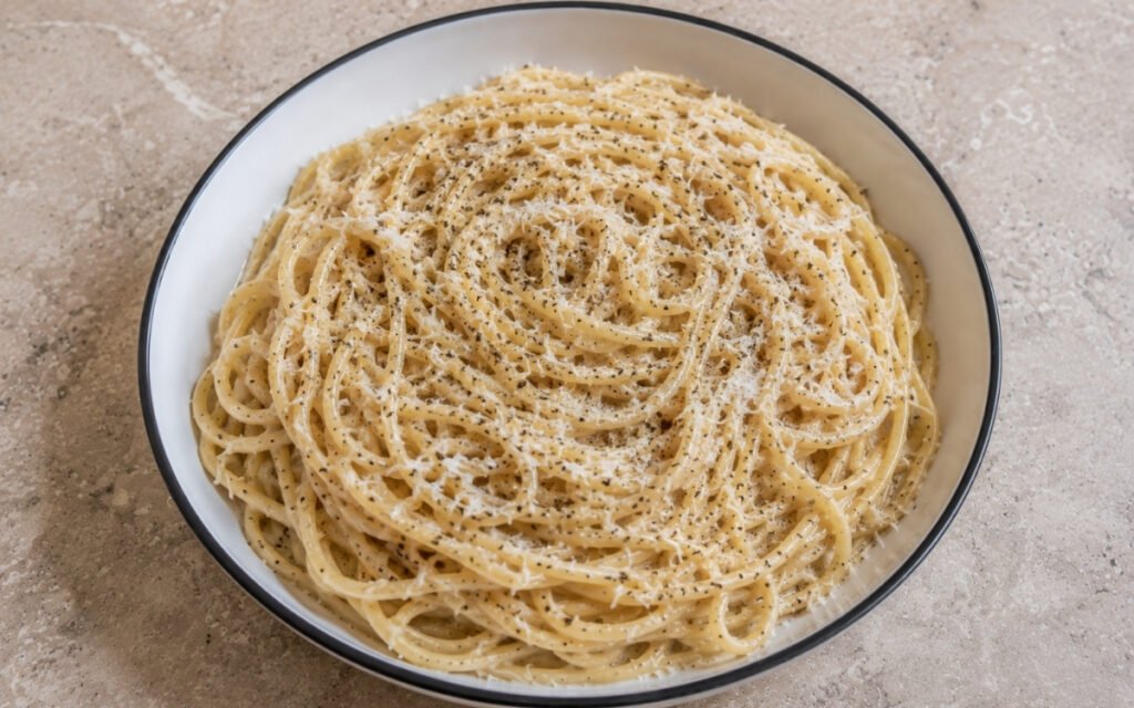 Classic cacio e pepe with tonnarelli in creamy pale Pecorino sauce with visible cracked black peppercorn flecks and additional grated Pecorino in a white ceramic bowl with black rim