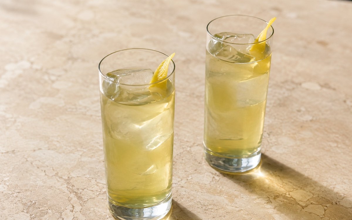 Cold brew green tea in a tall glass showing pale golden-green still drink over ice with a lemon peel twist on marble surface