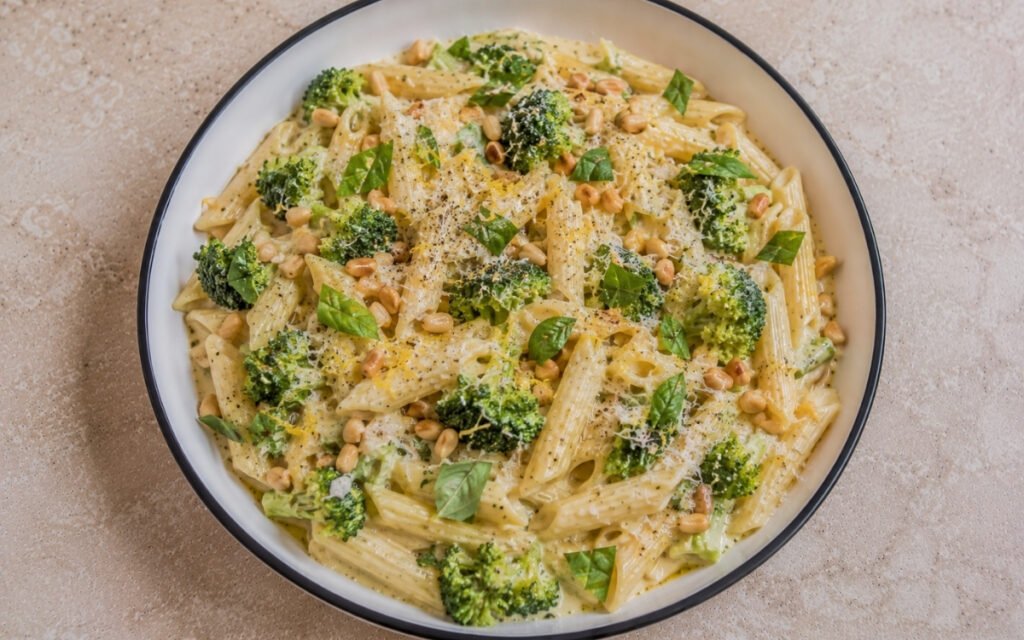Creamy broccoli lemon pasta with penne in pale golden mascarpone-Parmigiano sauce, bright green broccoli florets, lemon zest, torn basil and toasted pine nuts in a white ceramic bowl with black rim