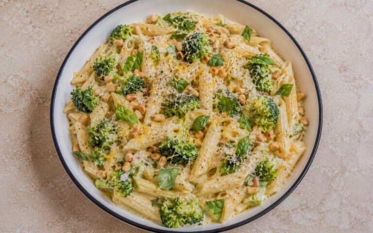 Creamy broccoli lemon pasta with penne in pale golden mascarpone-Parmigiano sauce, bright green broccoli florets, lemon zest, torn basil and toasted pine nuts in a white ceramic bowl with black rim