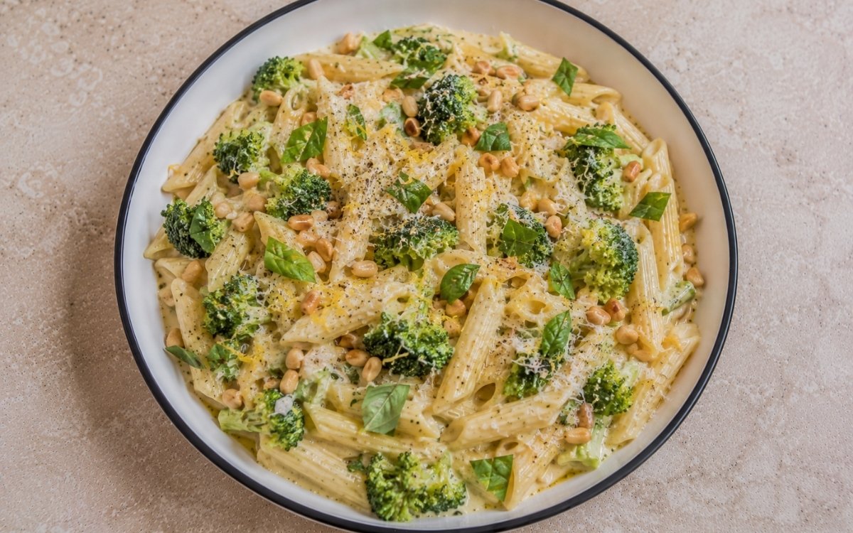Creamy broccoli lemon pasta with penne in pale golden mascarpone-Parmigiano sauce, bright green broccoli florets, lemon zest, torn basil and toasted pine nuts in a white ceramic bowl with black rim