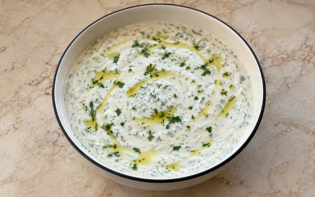 Bowl of creamy white yogurt sauce with green herbs and lemon zest on marble surface