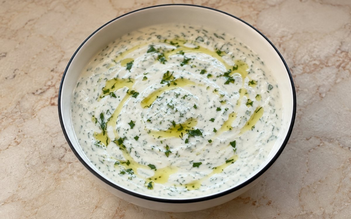Bowl of creamy white yogurt sauce with green herbs and lemon zest on marble surface