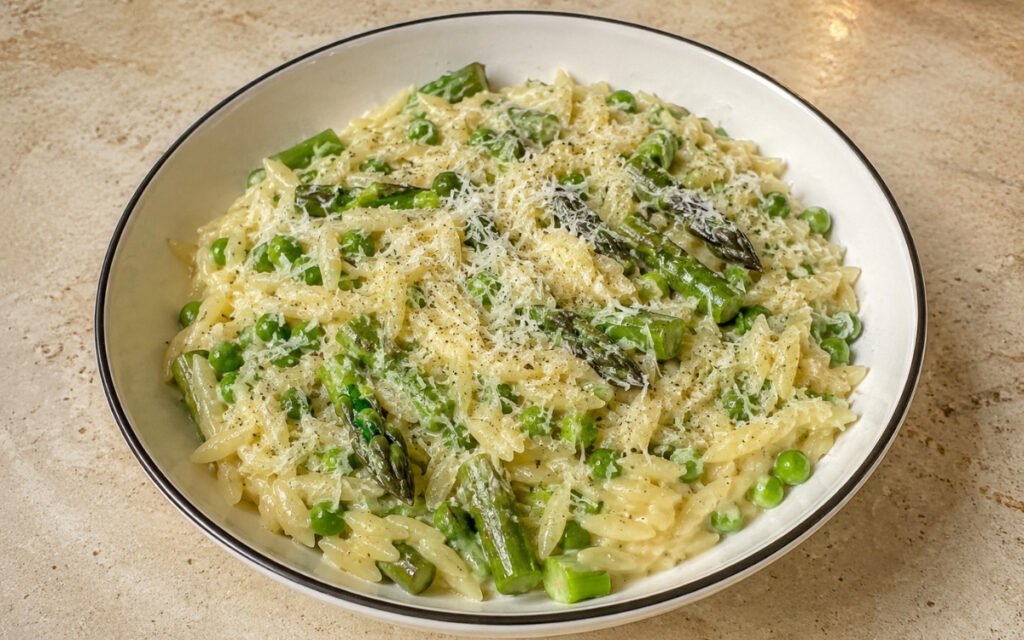 Creamy pea and asparagus orzo in a wide white bowl showing vibrant green sauce coating orzo with seared asparagus pieces and Parmigiano Reggiano scattered on top on marble surface