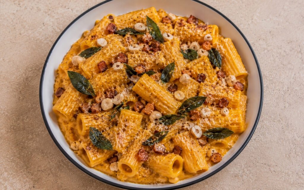 Creamy pumpkin pasta with rigatoni in golden-orange pumpkin cream sauce, crispy pancetta, crispy fried sage leaves, toasted hazelnuts and Parmigiano-Reggiano in a white ceramic bowl with black rim
