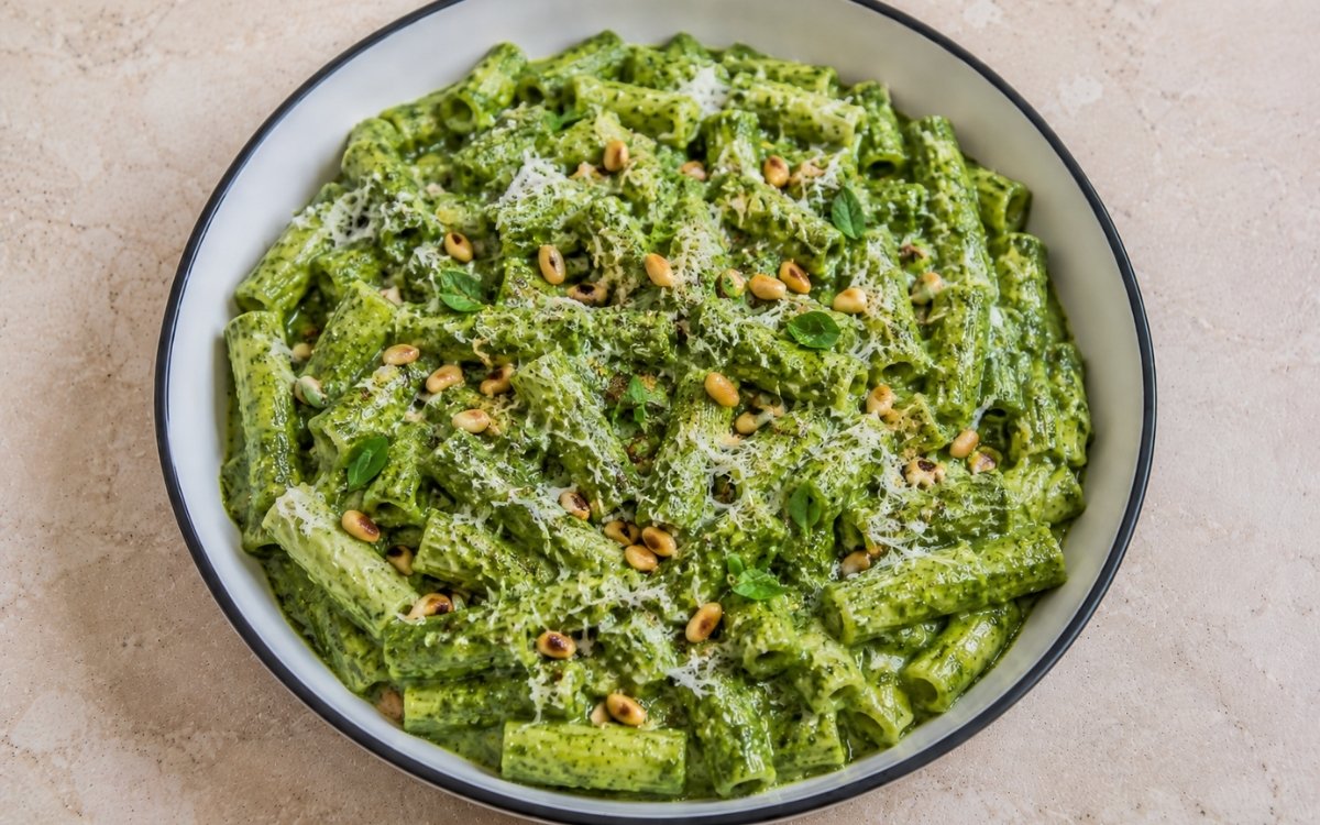 Creamy spinach and herb green sauce pasta with rigatoni in vivid green blended spinach, basil, parsley and mint cream sauce with toasted pine nuts and Parmesan in a white ceramic bowl with black rim