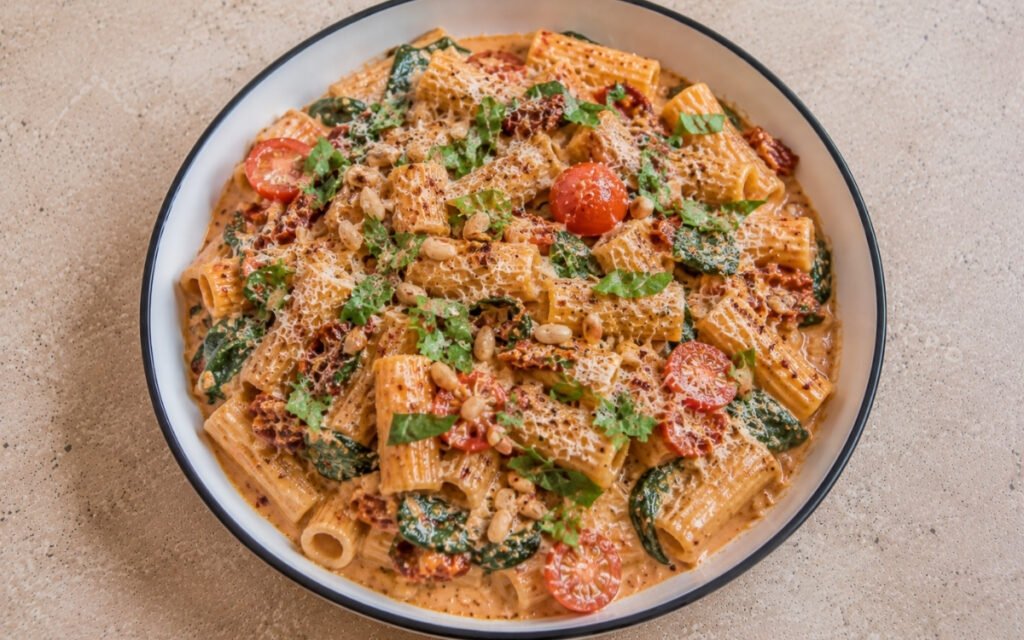 Creamy sun-dried tomato pasta with rigatoni in pale orange-red cream sauce with sun-dried tomatoes, halved cherry tomatoes, wilted spinach, torn basil, toasted pine nuts and Parmesan in a white ceramic bowl with black rim