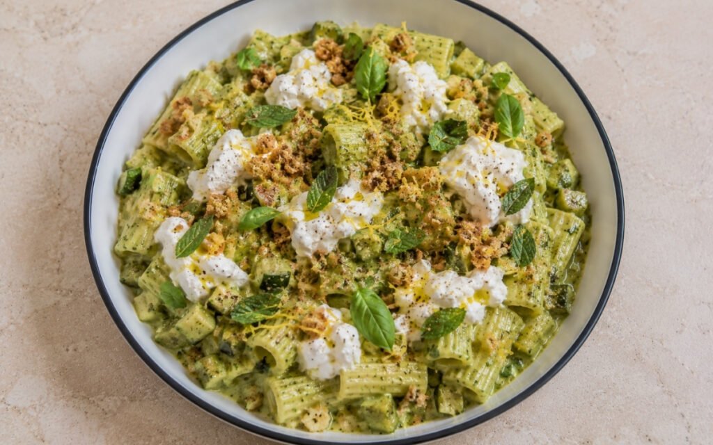 Creamy zucchini pasta with rigatoni in vibrant green zucchini cream sauce, torn burrata pieces with spilling cream, golden lemon breadcrumbs, fresh basil and mint in a white ceramic bowl with black rim