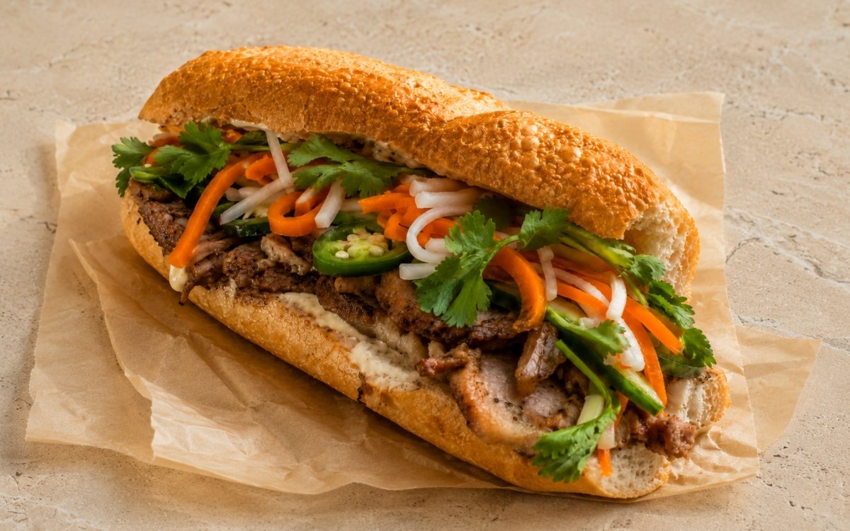 Crispy pork bánh mì sandwich showing crackling pork belly with pickled carrot and daikon, cucumber, and fresh cilantro in a crusty baguette on marble surface