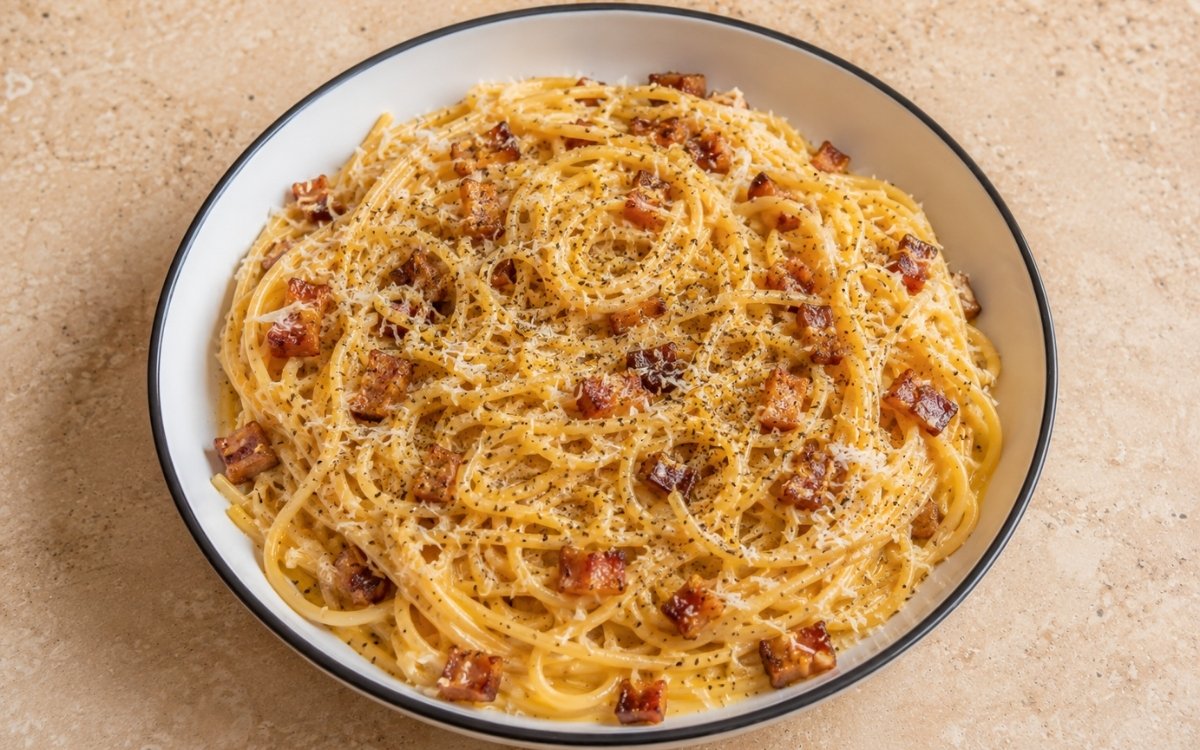 Authentic carbonara pasta with spaghetti in pale yellow egg-yolk sauce, crispy golden guanciale lardons, generous Pecorino Romano and freshly cracked black pepper in a white ceramic bowl with black rim