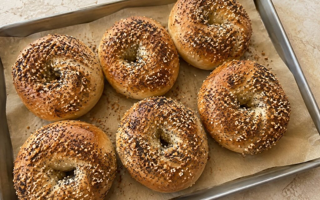 Everything bagels on a wire cooling rack showing deep golden crust with everything seasoning coating of sesame seeds, poppy seeds, dried onion, and garlic on marble surface