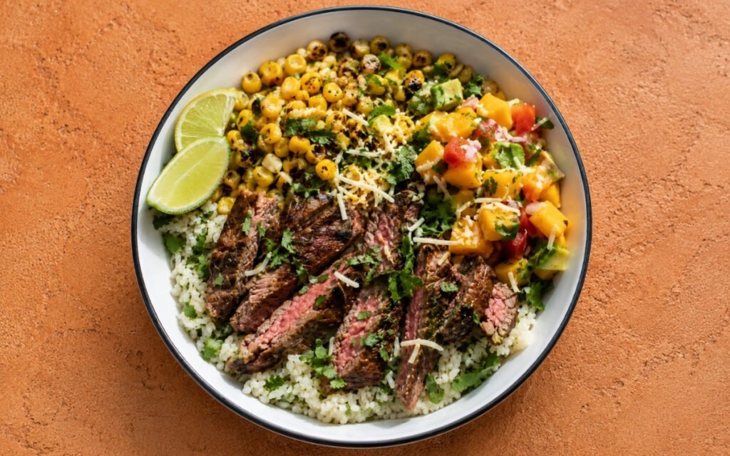 Overhead view of flank steak and mango salsa rice bowl in a ceramic bowl with charred corn, avocado mango salsa, shredded cheese, and lime wedges on a grey marble surface