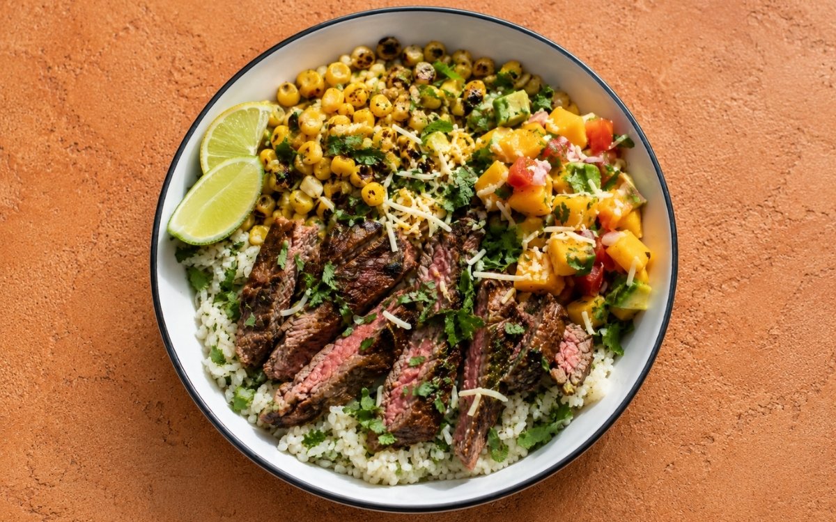 Overhead view of flank steak and mango salsa rice bowl in a ceramic bowl with charred corn, avocado mango salsa, shredded cheese, and lime wedges on a grey marble surface