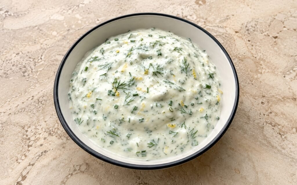 Fresh dill yogurt sauce in a white bowl with fresh dill fronds and lemon on white marble