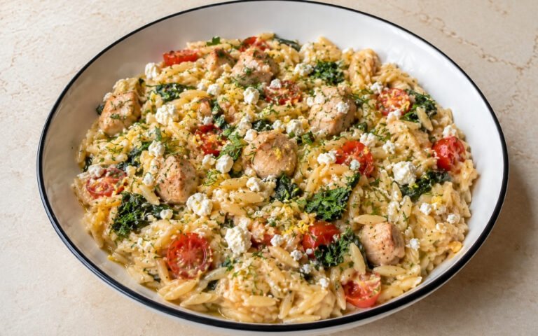 Fresh lemon chicken orzo with creamy orzo pasta, diced chicken thighs, halved cherry tomatoes, wilted spinach, crumbled feta, fresh dill and parsley in a white ceramic bowl with black rim